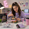 A young woman with her hair in a ponytail is smiling and assembling a colorful brick-built cafe model on a desk. The scene is set in a cozy, decorated room with a pink neon sign, shelves, and various stationery items.