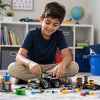 A young boy with dark hair sits cross-legged on a carpeted floor in a child's playroom, building a black and gray toy vehicle with small LEGO minifigures and loose bricks. LEGO branding is visible on the vehicle and scattered pieces.