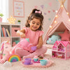 A young girl with a pink bow in her hair sits on a carpeted floor in a child's bedroom, playing with a pink toy tea set and cupcake cups on a tray. She is wearing a pink dress and pink leggings.